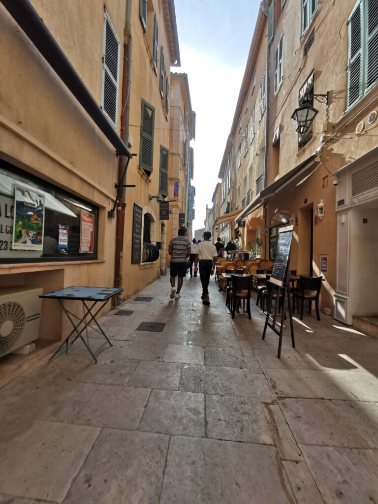 Narrow cobbled street in St Tropez lined with outdoor café tables, menu boards and warm pastel buildings on the French Riviera.