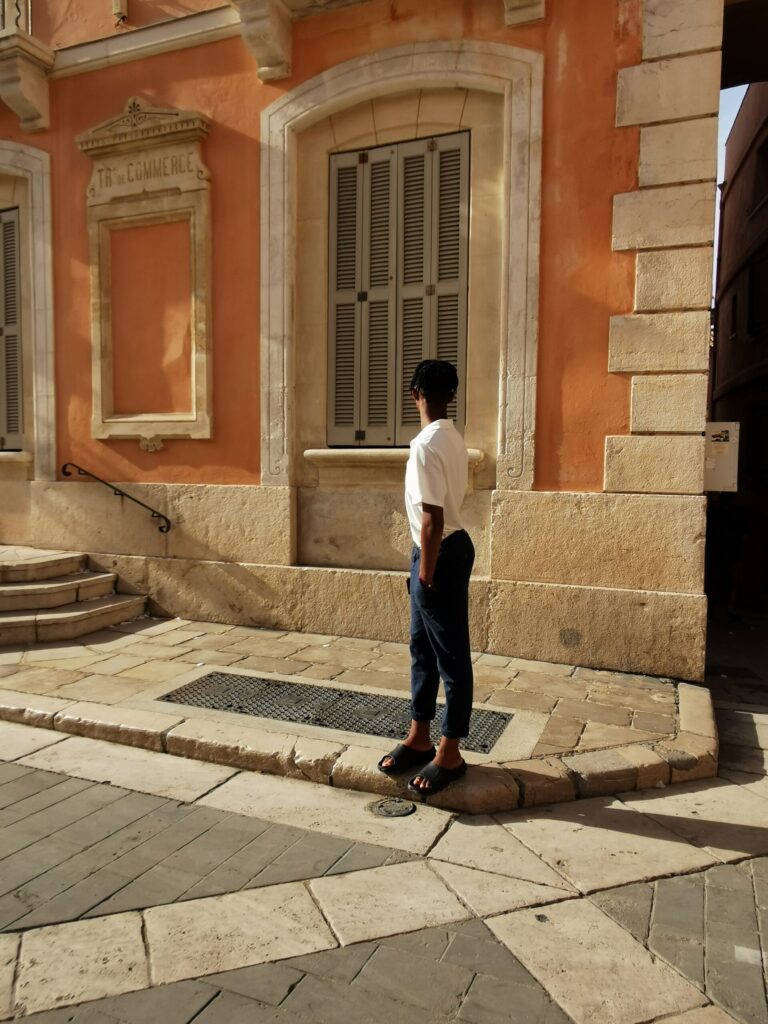 Teenager standing beside a warm peach coloured building with traditional shutters in Saint Tropez old town on the French Riviera.