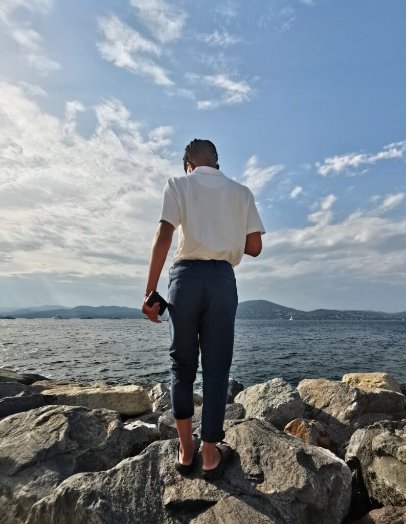 Boy standing on seaside rocks looking out across the Mediterranean Sea towards the Saint Tropez coastline under a wide blue sky.