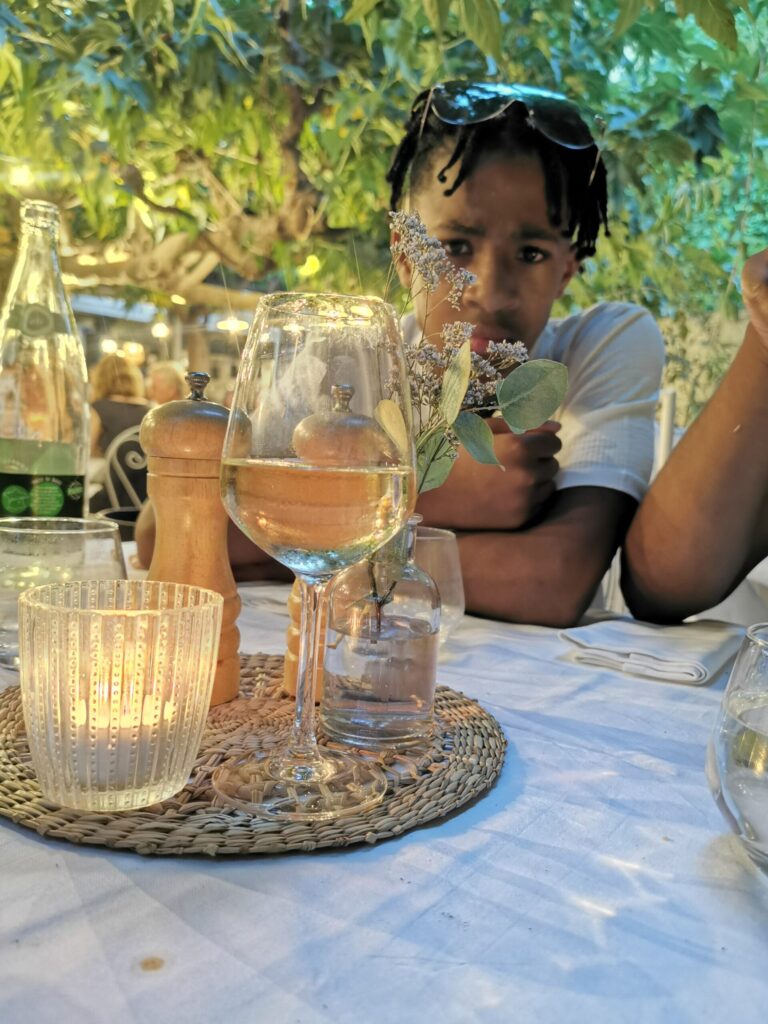 Teenager sitting at an outdoor terrace table beneath leafy trees at La Ramade restaurant in St Tropez.