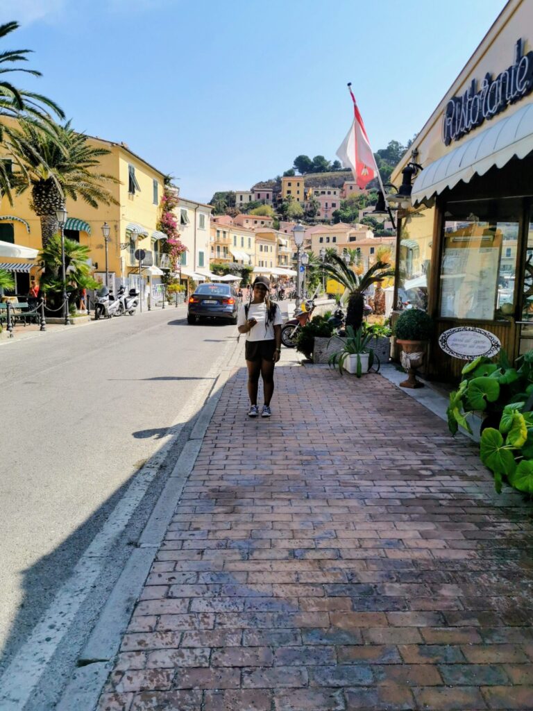 Teenage girl standing in a colourful European coastal village street with pastel buildings. 