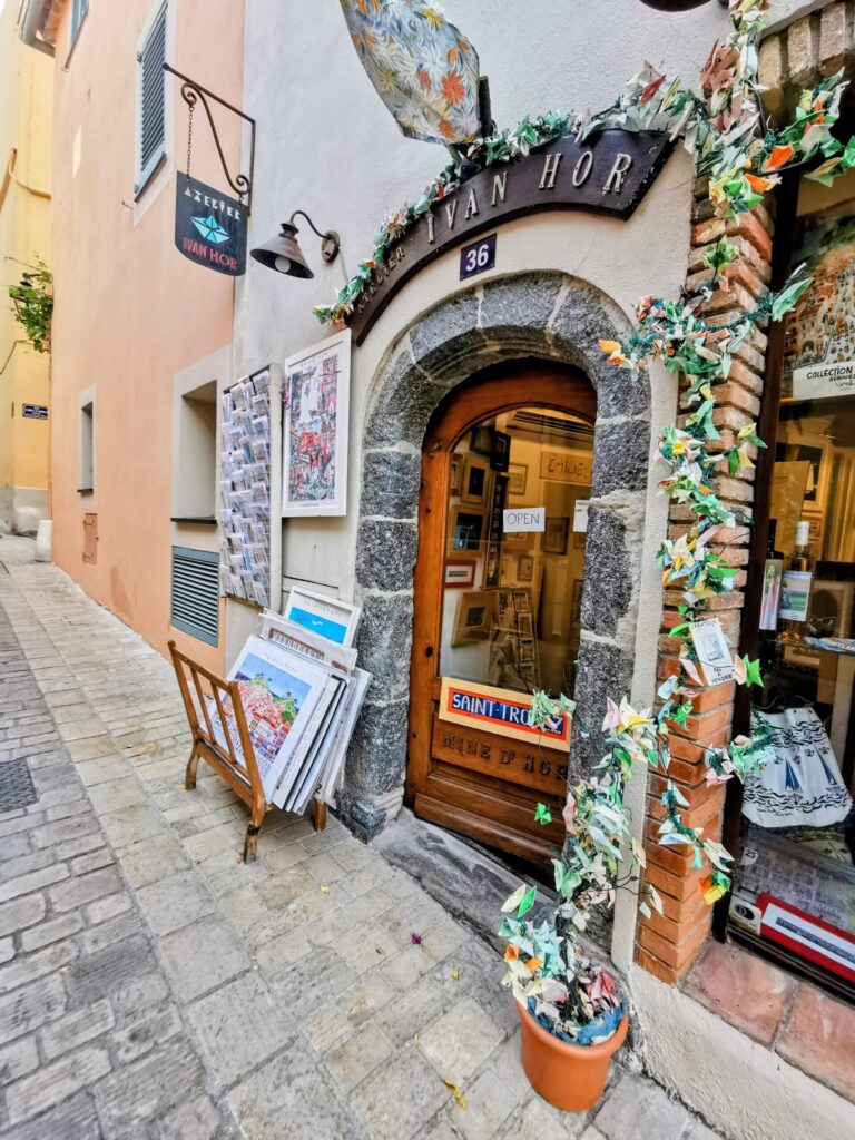 Independent bookshop tucked away in a narrow Saint - Tropez side street, with artwork displayed outside and colourful paper decorations framing the entrance, reflecting the quieter, creative side of town.
