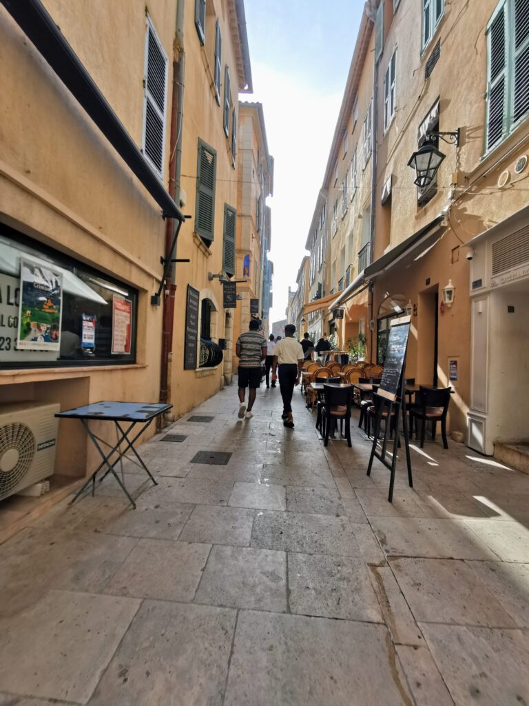 Sunlit cobbled street in Saint-Tropez, lined with pastel buildings, cafes, terraces and local shops, capturing the town's quieter side away from the marina. 