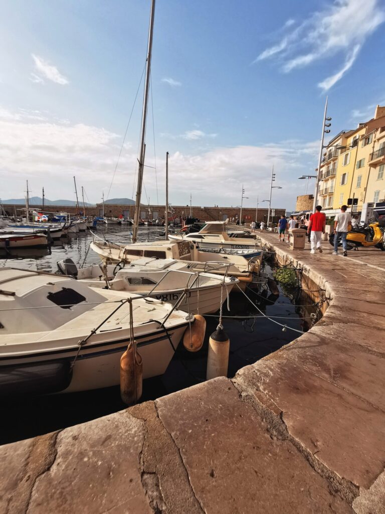 Snall fishing boats moored along the Saint - Tropez, with people strolling beside the harbour under the late afternoon sun. 