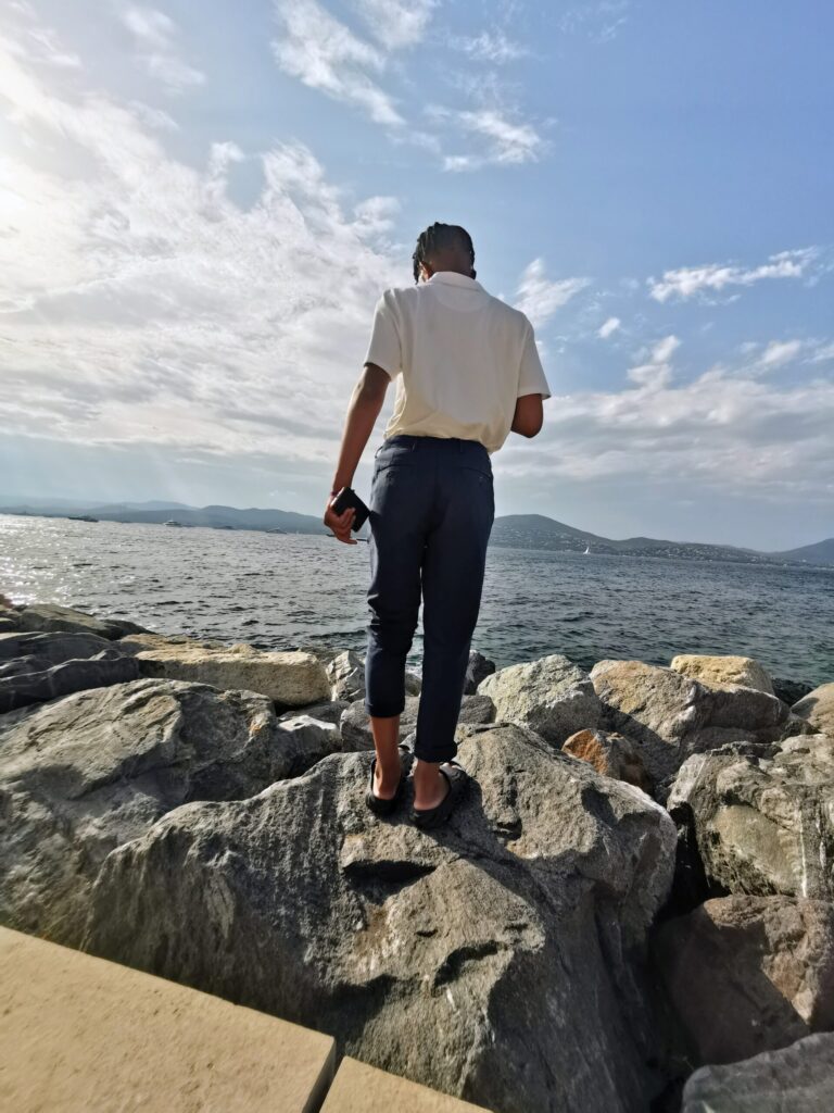 Young man standing on large rocks beside the sea in Saint - Tropez, looking out across the water towards the distant coastline under a bright summer sky.