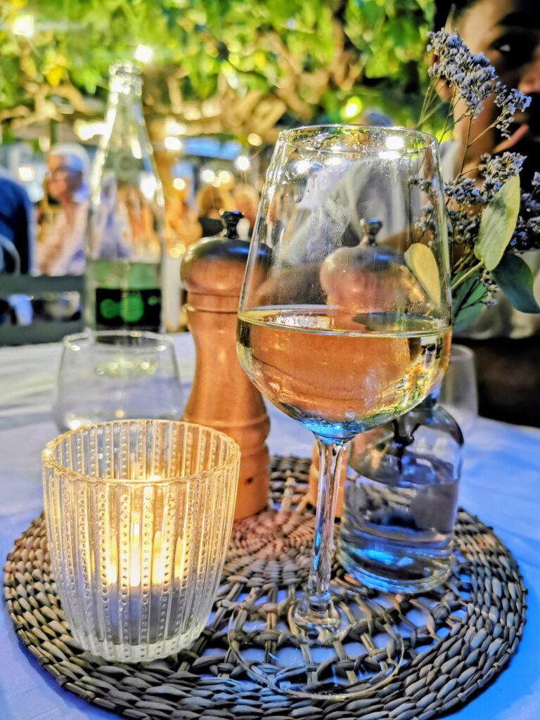Glass of white wine and candlelit table at an outdoor restaurant in Saint - Tropez, surrounded by soft lighting and greenery on a warm summer evening. 