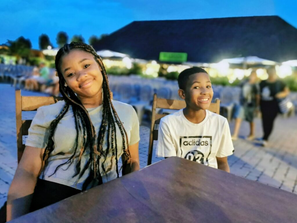 Two children sitting together at an outdoor restaurant in Italy during a warm summer evening, enjoying a relaxed family holiday. 