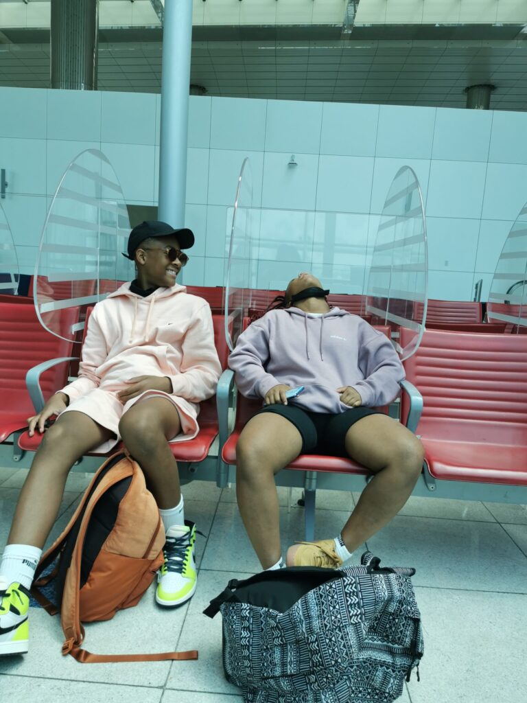 Young woman sleeping at the airport whilst her brother laughs besides her.